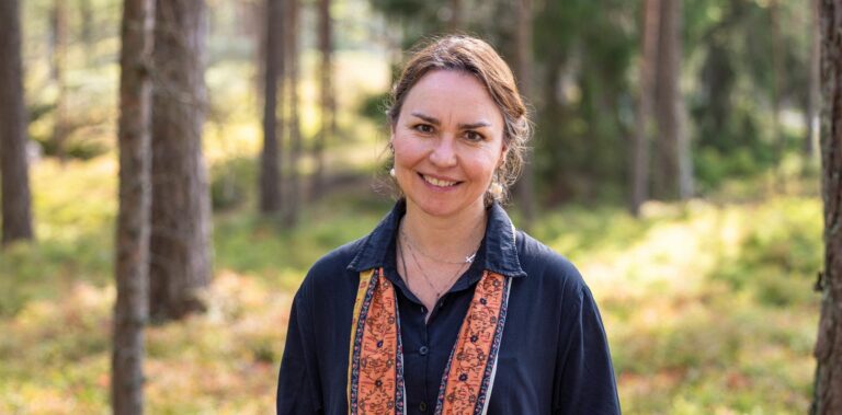Nainen seisoo mäntymetsässä, katsoo kameraan ja hymyilee