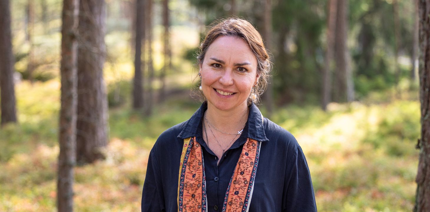 Nainen seisoo mäntymetsässä, katsoo kameraan ja hymyilee