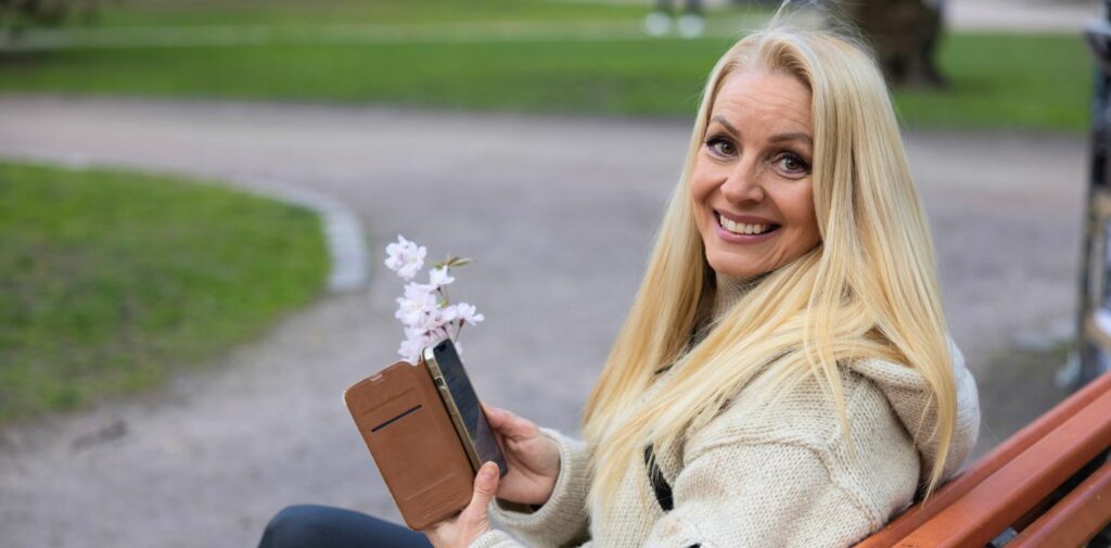 Nainen istuu penkillä puhelin kädessään ja katsoo hymyillen kameraan