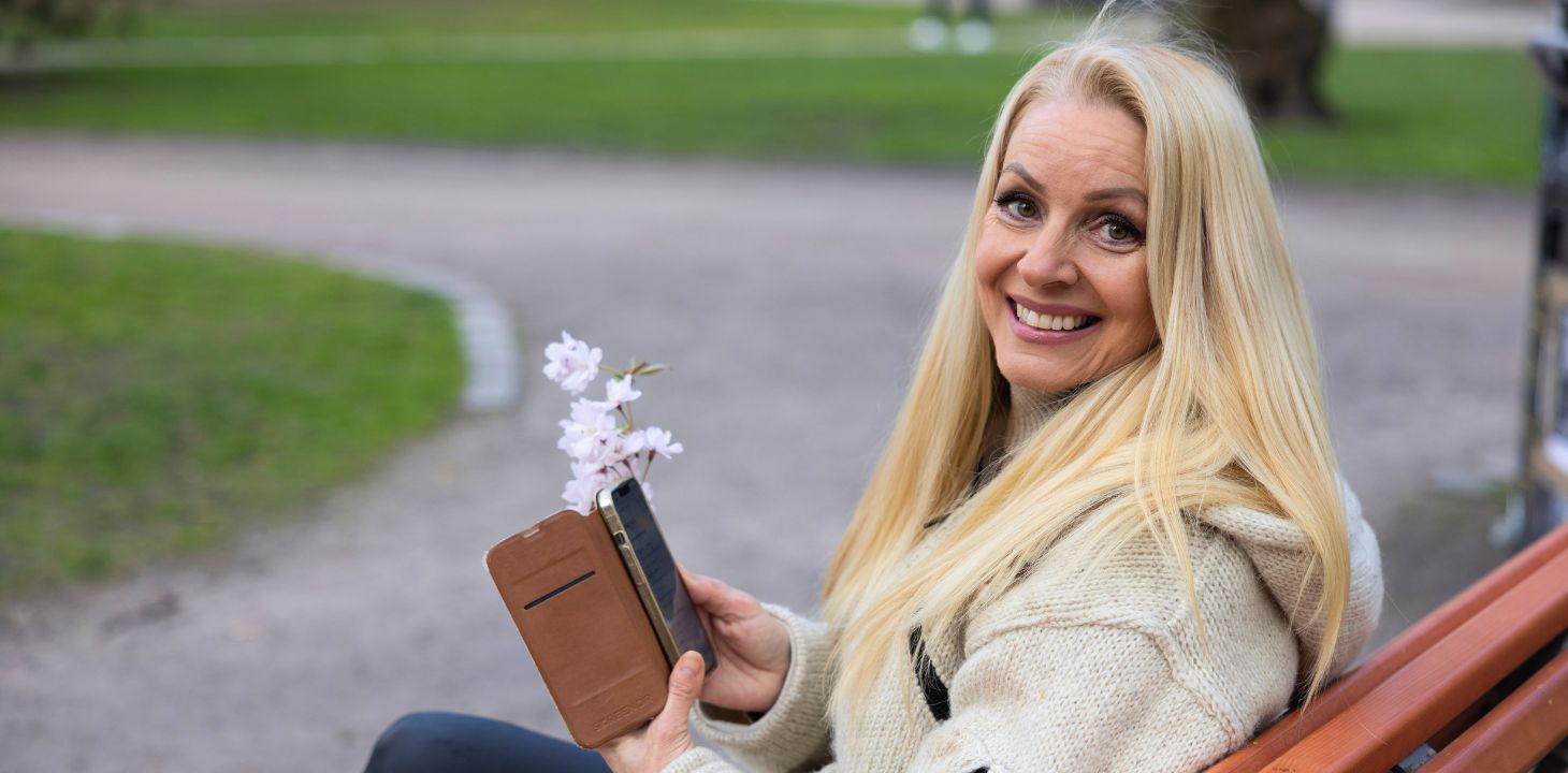 Nainen istuu penkillä puhelin kädessään ja katsoo hymyillen kameraan