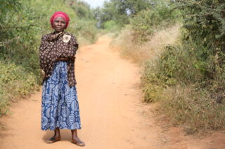 Tansanialaisen kylän naispuolinen kylänjohtaja seisoo hiekkaisella kylätiellä itsevarmassa asennossa.