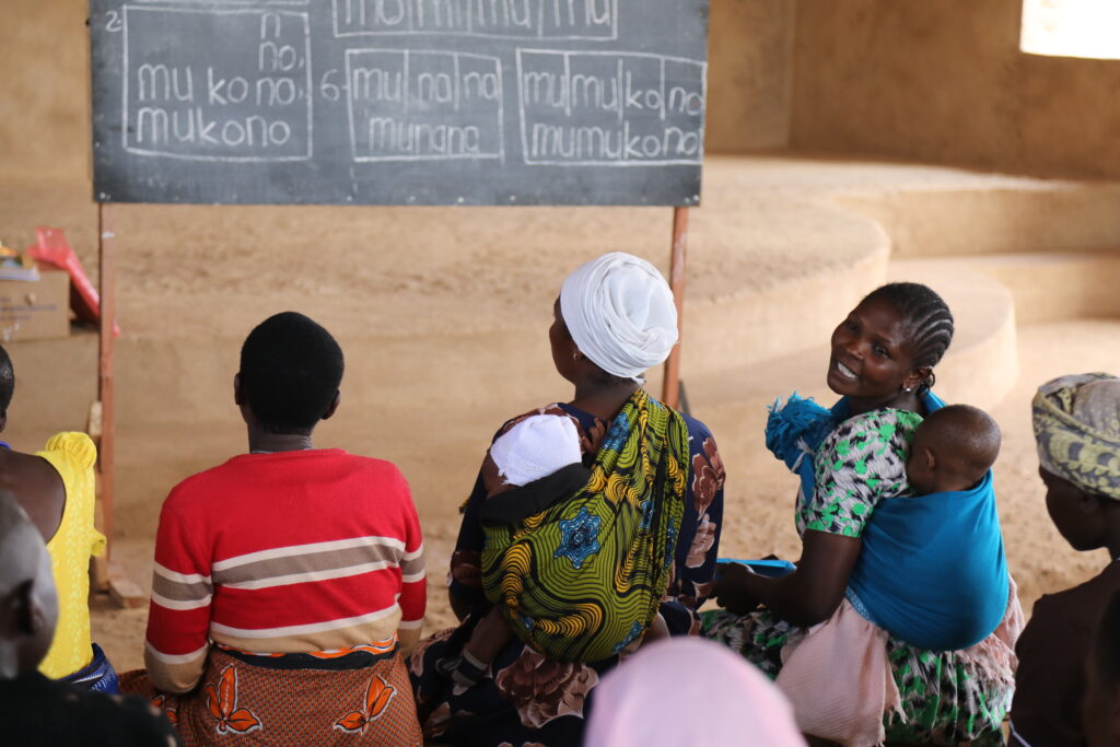 Tansanialaisen lukutaitoluokan naisia istumassa liitutaulun edessä. Useilla naisilla on pieni lapsi kääriittynä liinaan ja kiinnitettynä selkään.