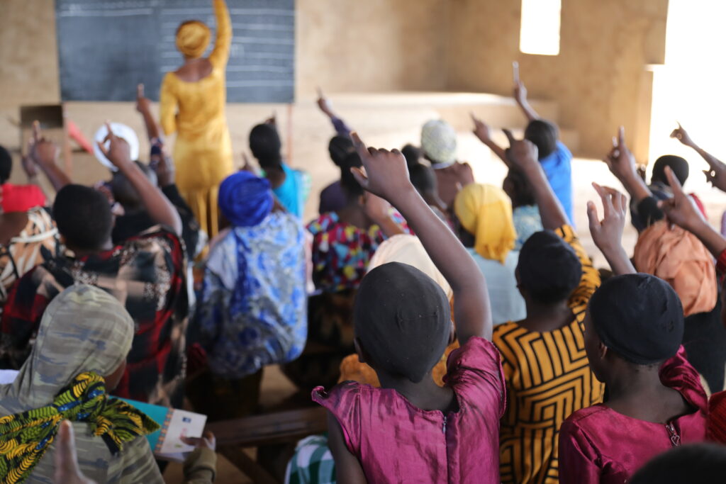 Paljon tnsanialaisen lukutaitoluokan naisia opetuksen tuoksinassa viittamaamassa innokkaasti. Taustalla näkyy opettaja kirjoittamassa liitutaululle.