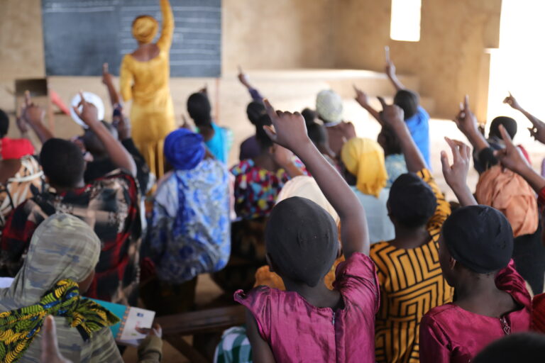 Paljon tnsanialaisen lukutaitoluokan naisia opetuksen tuoksinassa viittamaamassa innokkaasti. Taustalla näkyy opettaja kirjoittamassa liitutaululle.