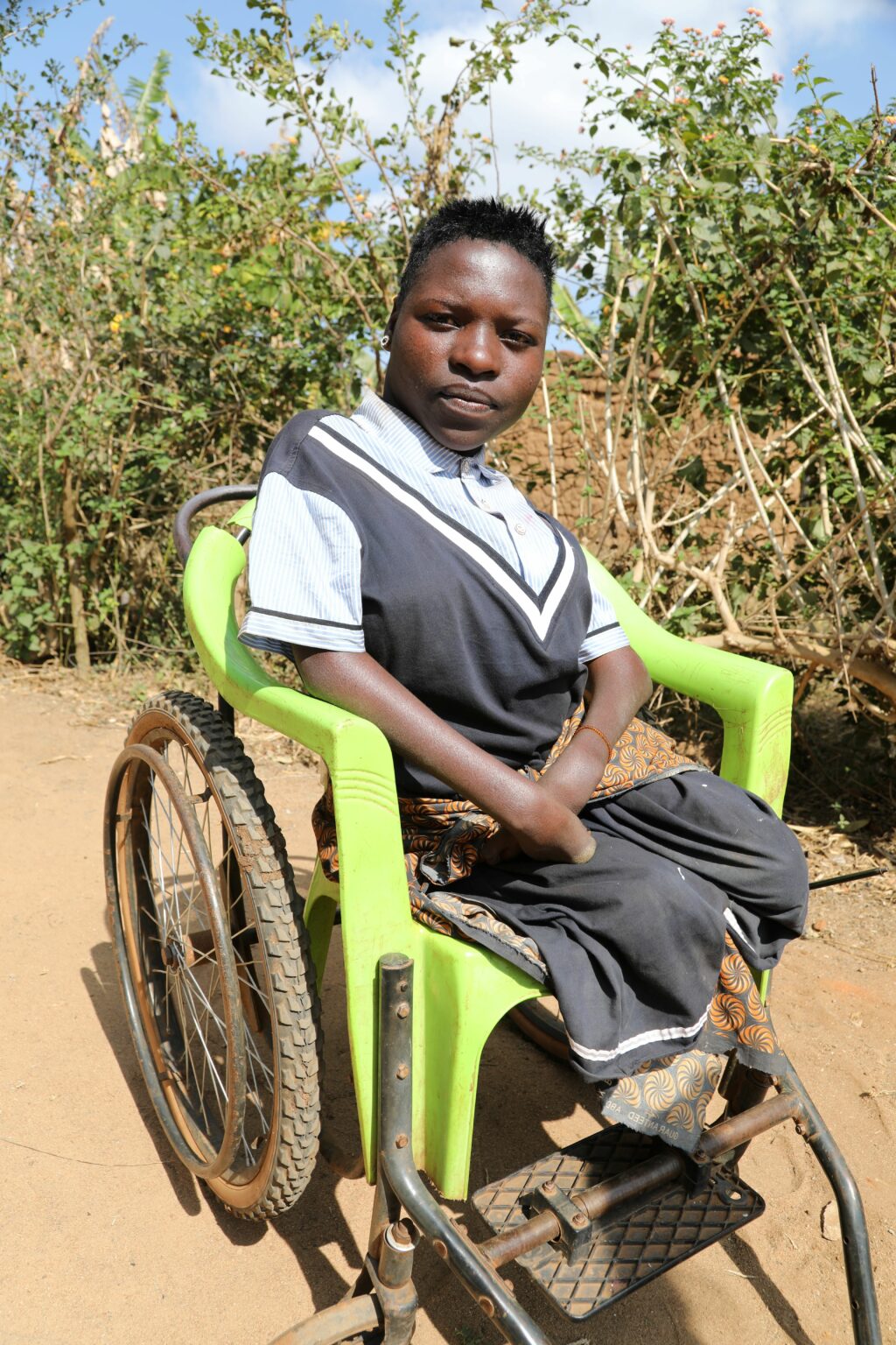 Liikuntavaimmainen tansanialainen lukutaito-opiskelija istuu pyörätuolissa ulkona. Taustalla pensaita.