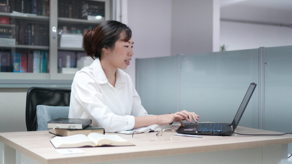 Thaimaalainen nainen käyttää tietokonetta toimistoympäristössä, pöydällä kirjoja ja Raamattu.