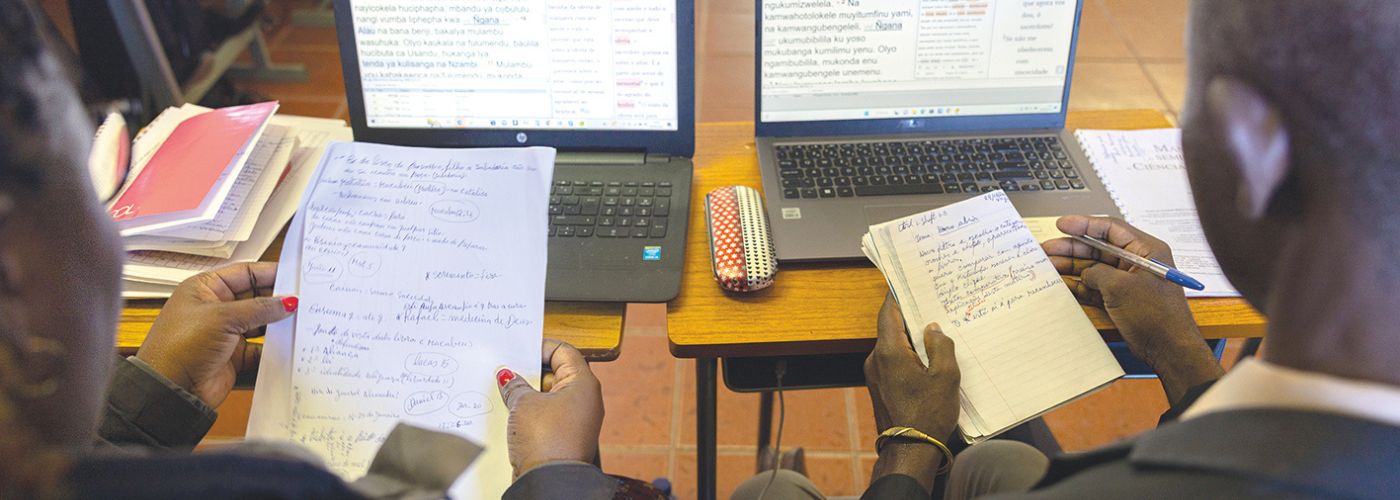 takaapäin kuva kahdesta raamatunkääntäjästä, heillä käsissään muistiinpanot ja edessä tietokone