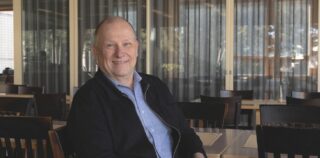 Mies istuu kahvilassa, katsoo kameraan ja hymyilee